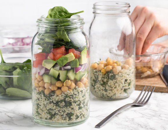 Two jars of quinoa with chickpeas, one filled with vegetables, and one being filled